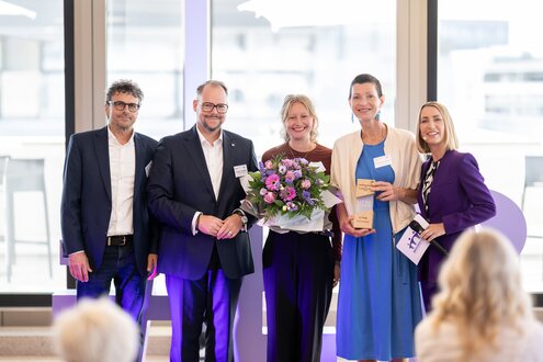 Fünf Personen stehen nebeneinander bei der Preisverleihung. In der Mitte stehen die Preisträgerinnen mit einer Trophäe und einem Blumenstrauß. Links stehen der Laudator und ein Vorstandmitglied der Evangelischen Bank. Rechts steht die Moderatorin der Preisverleihung.